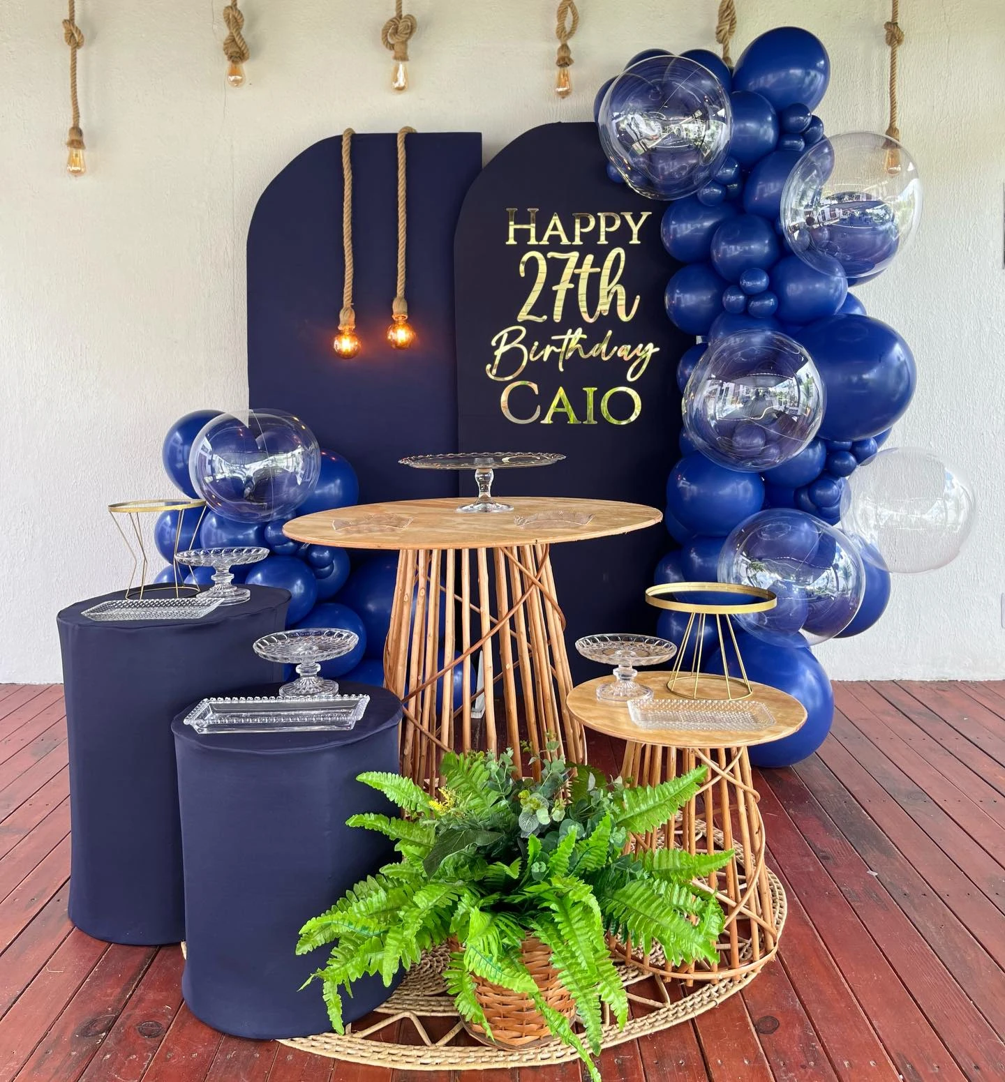 Aniversário de 27 anos com painéis azuis, balões metalizados, mesas de madeira e samambaias.