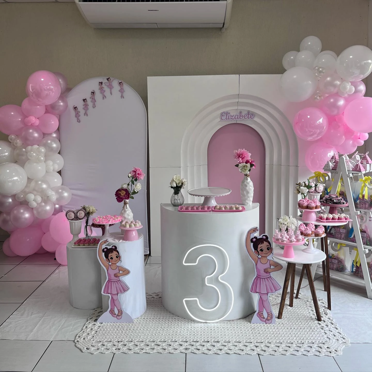 Decoração infantil de bailarina com arcos brancos, balões rosa e mesas de doces.