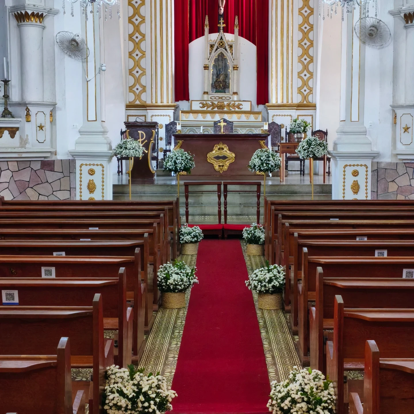 Corredor de igreja preparado para casamento com passadeira vermelha e arranjos de flores brancas.