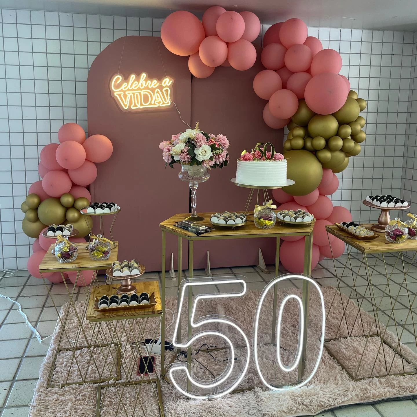 Festa de 50 anos com painel rosado, balões rosa e verde-oliva e mesas de doces.