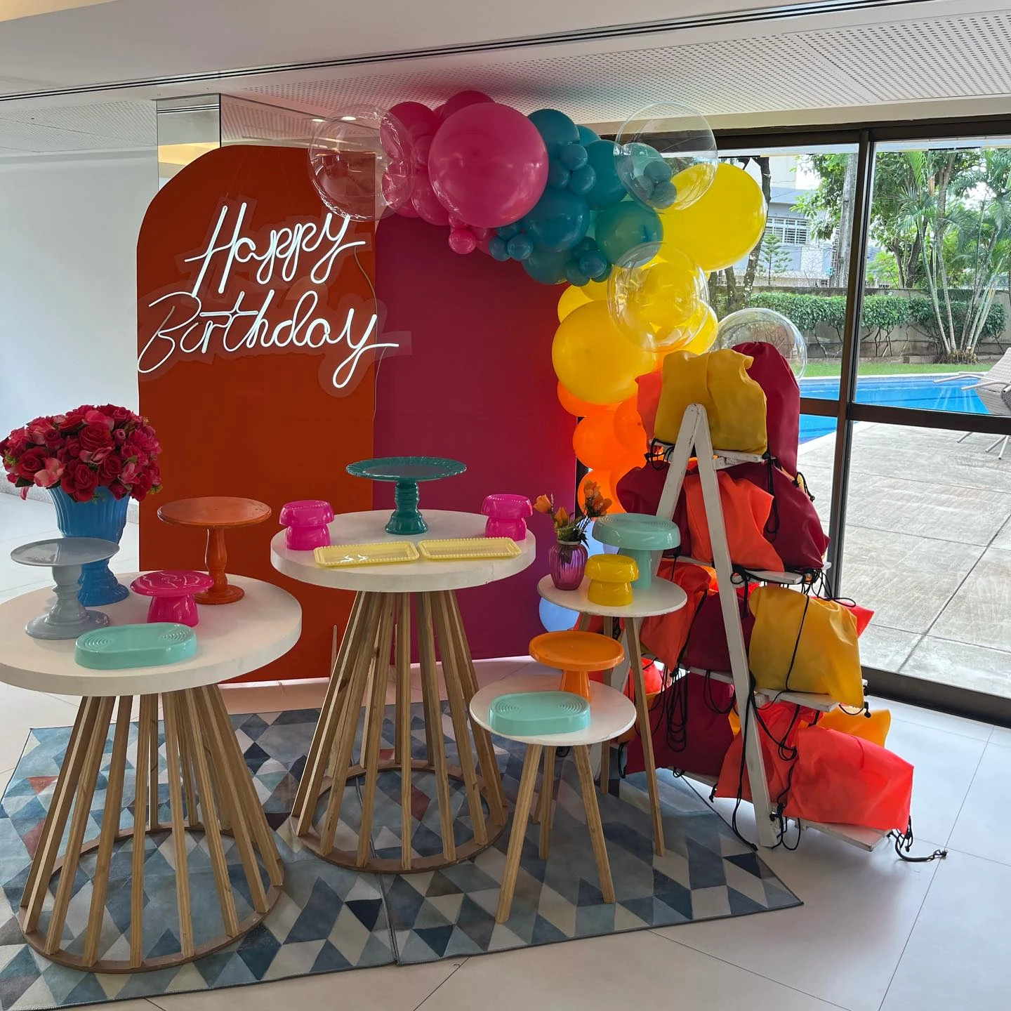 Decoração pool party com painéis laranja e rosa, balões amarelos e azuis e mesas coloridas.